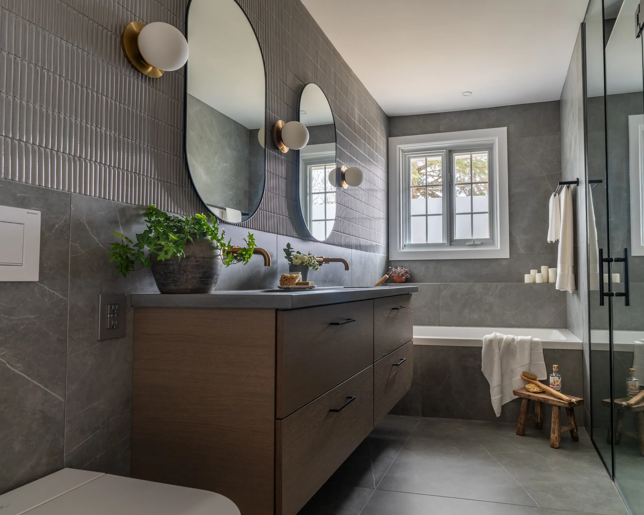 Primary bathroom in Boucherville — walnut floating vanity, aged-brass fixtures, microcement walls and oval mirrors.
