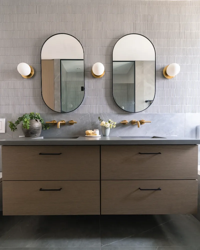 Walnut floating vanity head-on — twin oval mirrors, brass wall-mount faucets, fluted tile backdrop, Boucherville.